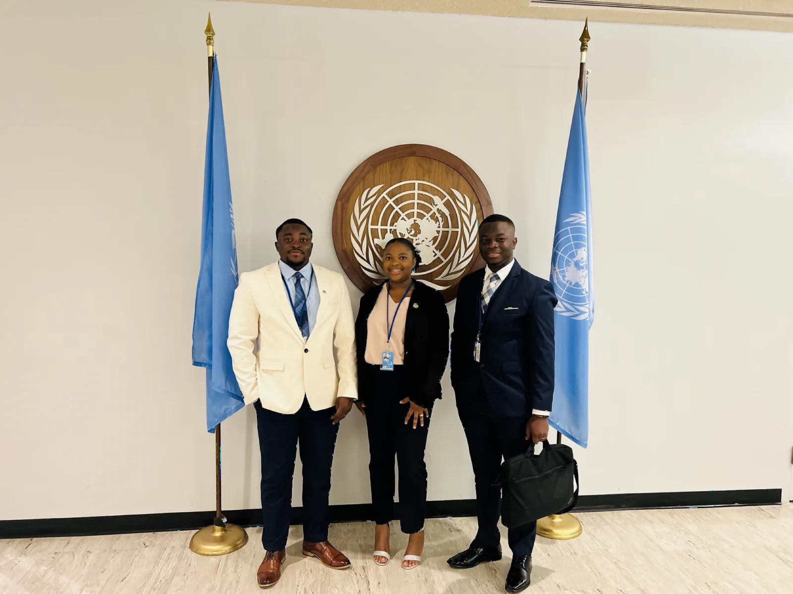 Three delegates standing in front of the United Nations emblem, between two United Nations flags.